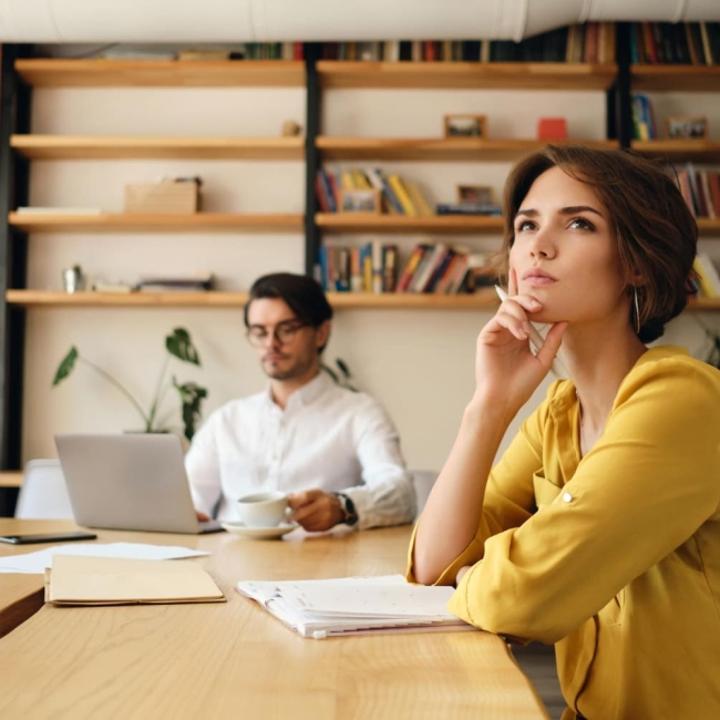 jeune-femme-pensive-assise-table-bloc-notes-regardant-reveusement-cote-tout-travaillant-collegue-arriere-plan-dans-bureau-moderne (1)