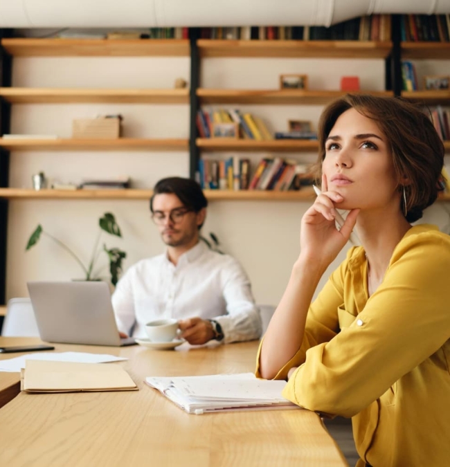 jeune-femme-pensive-assise-table-bloc-notes-regardant-reveusement-cote-tout-travaillant-collegue-arriere-plan-dans-bureau-moderne (1)