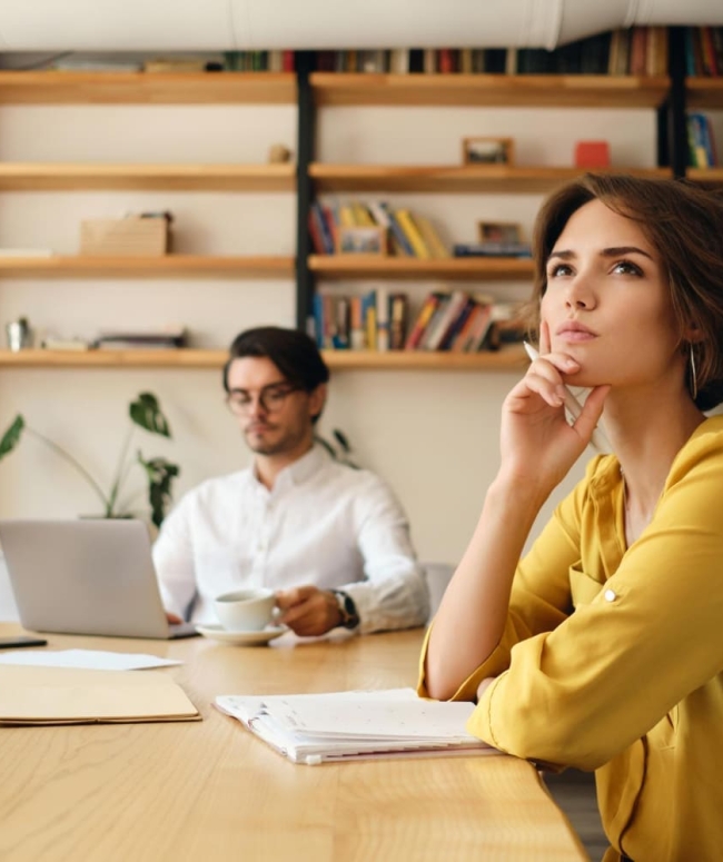 jeune-femme-pensive-assise-table-bloc-notes-regardant-reveusement-cote-tout-travaillant-collegue-arriere-plan-dans-bureau-moderne (1)